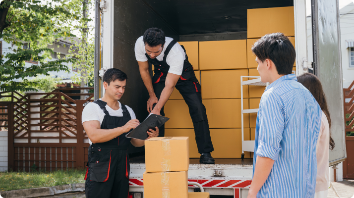 Two movers unload boxes from a truck, one checking a clipboard. A couple watches nearby. The scene conveys teamwork and organization.