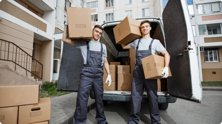 Two movers in uniforms stand smiling, each holding a box, in front of an open van loaded with boxes, set against an urban apartment building backdrop.
